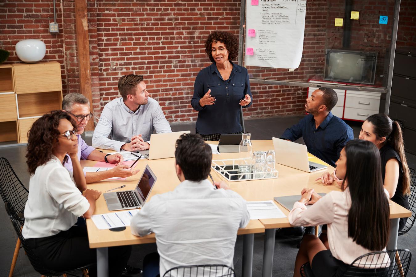Eight colleagues around a table in a brick office use a team meeting agenda template to address agenda items and talking points