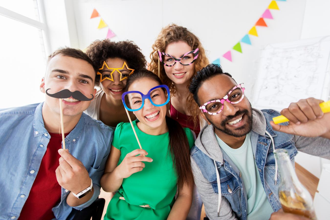 Five coworkers celebrate the birthday of someone in the office by dressing up and playing with silly props at a photo booth
