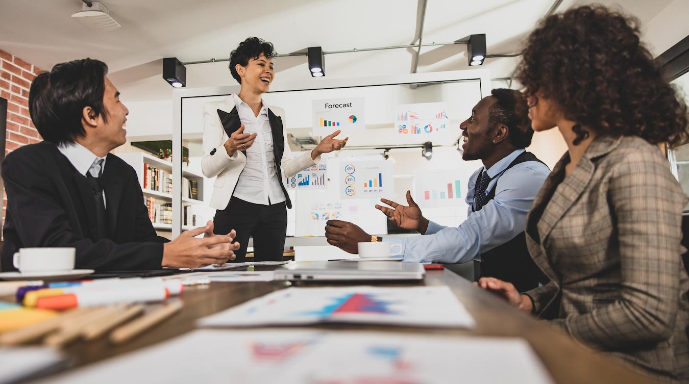 Four colleagues smile and greet each other in a conference room as they prepare for a mid year forecasting meeting at the office
