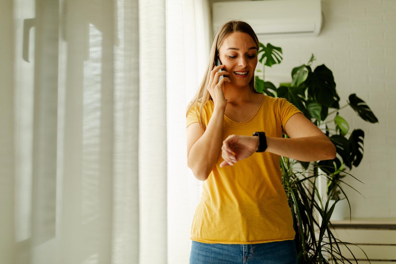 Person in a sunny room stands in front of a plant and looks at their watch because they are practicing their time management skills