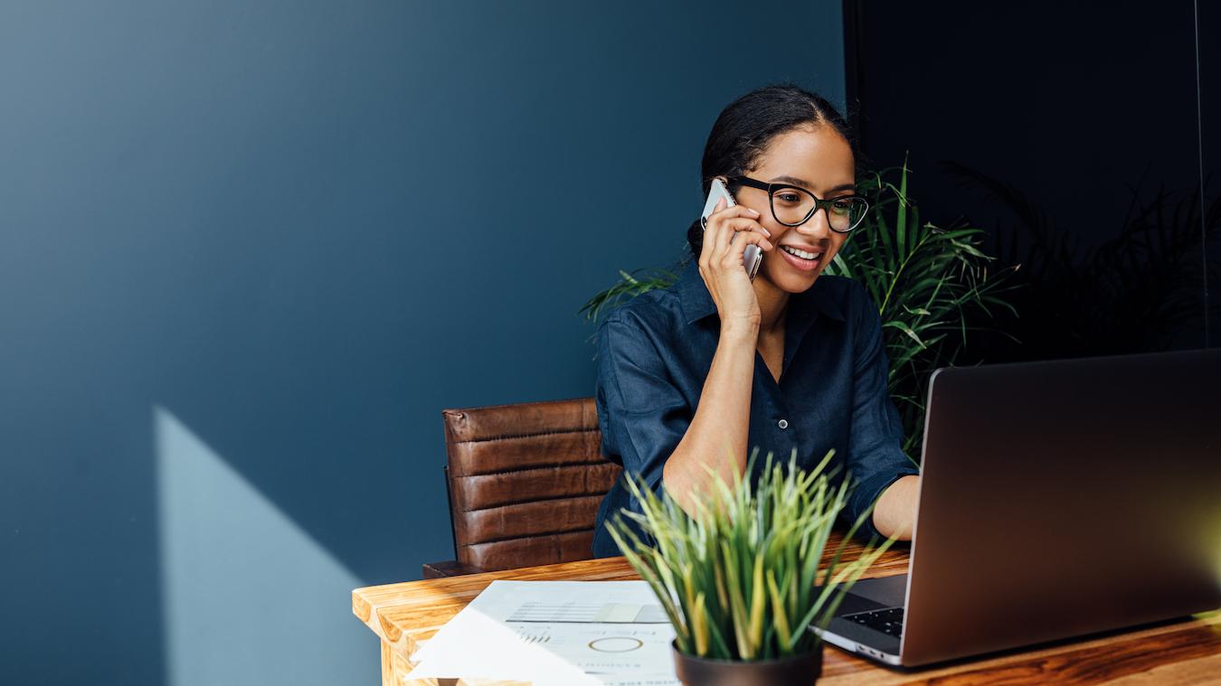Person in dark blue button up and glasses smiles as they talk on the phone and look at their computer
