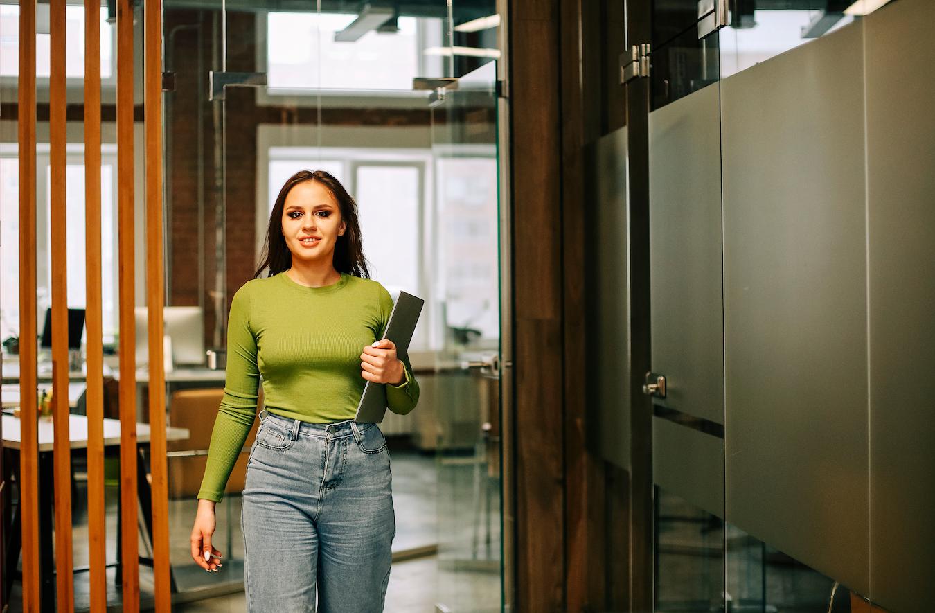 Person in green long sleeved shirt and light wash jeans leaves an office room carrying a laptop and looking calm happy and confident