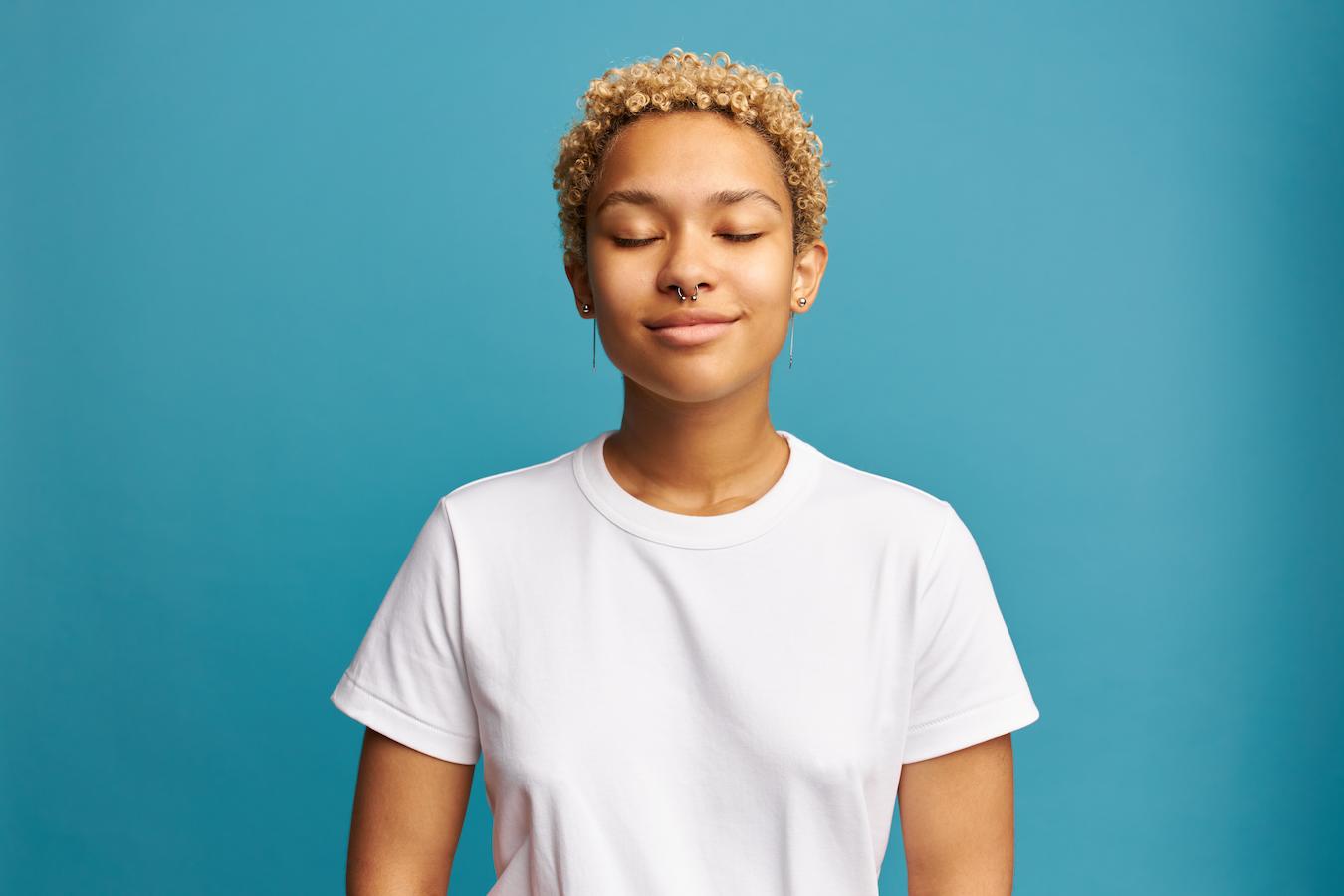 Person in white t shirt with blonde curls stands in front of a blue background and takes a deep breath to deal with a stressful moment in their life