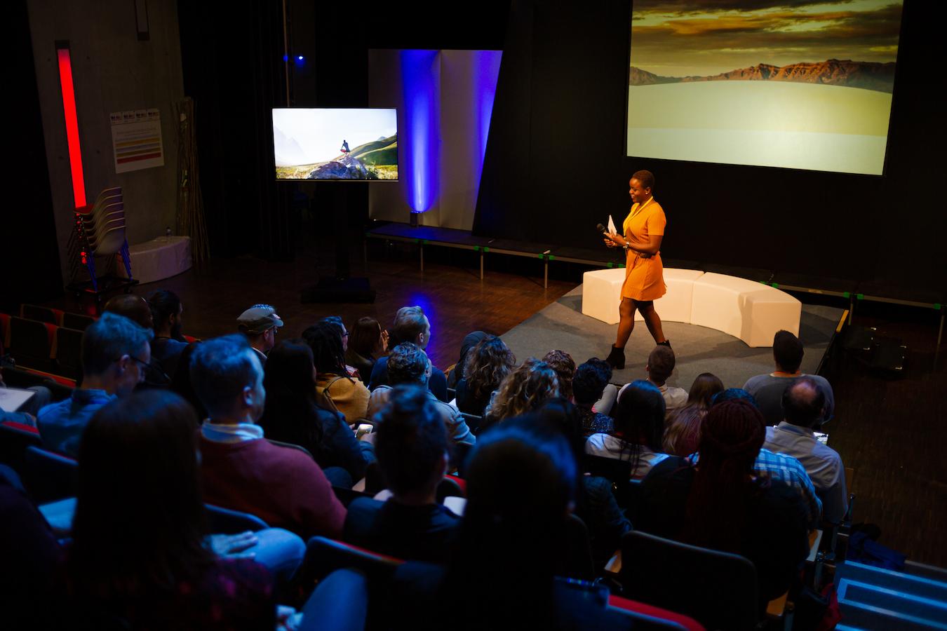 Person in yellow dress and black boots walks onstage in darkly lit theater to deliver a presentation with an important message