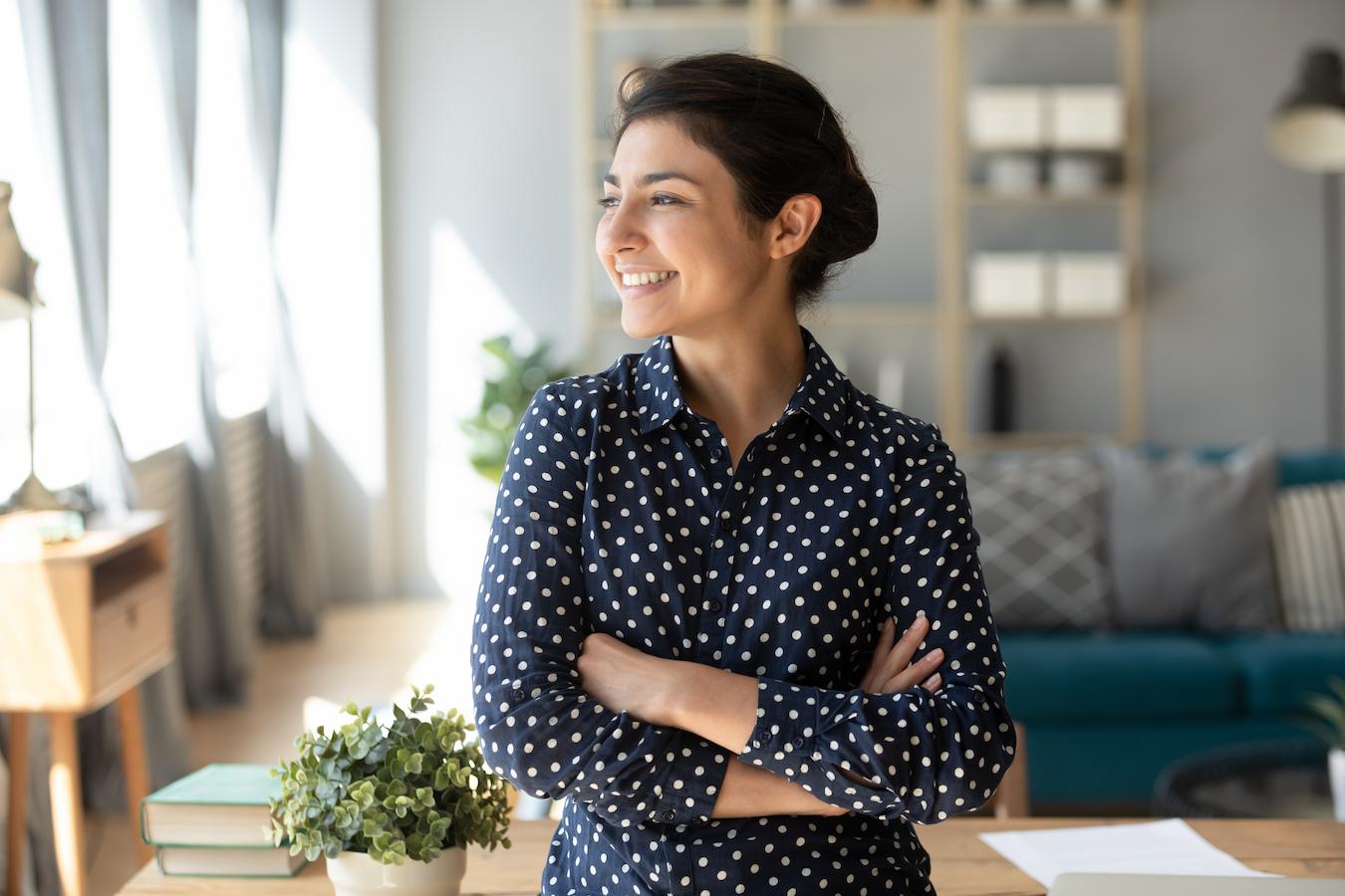 Person with brown hair in black and white polka dotted button down blouse folks arms across chest and looks toward the window of an office smiling happily
