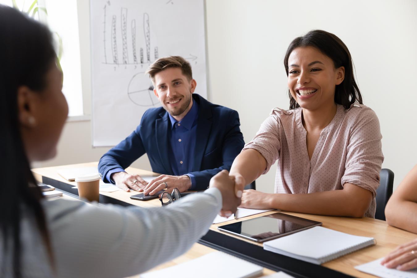 Two business colleagues shake hands and smile while another colleague sits next to them in a bright white office