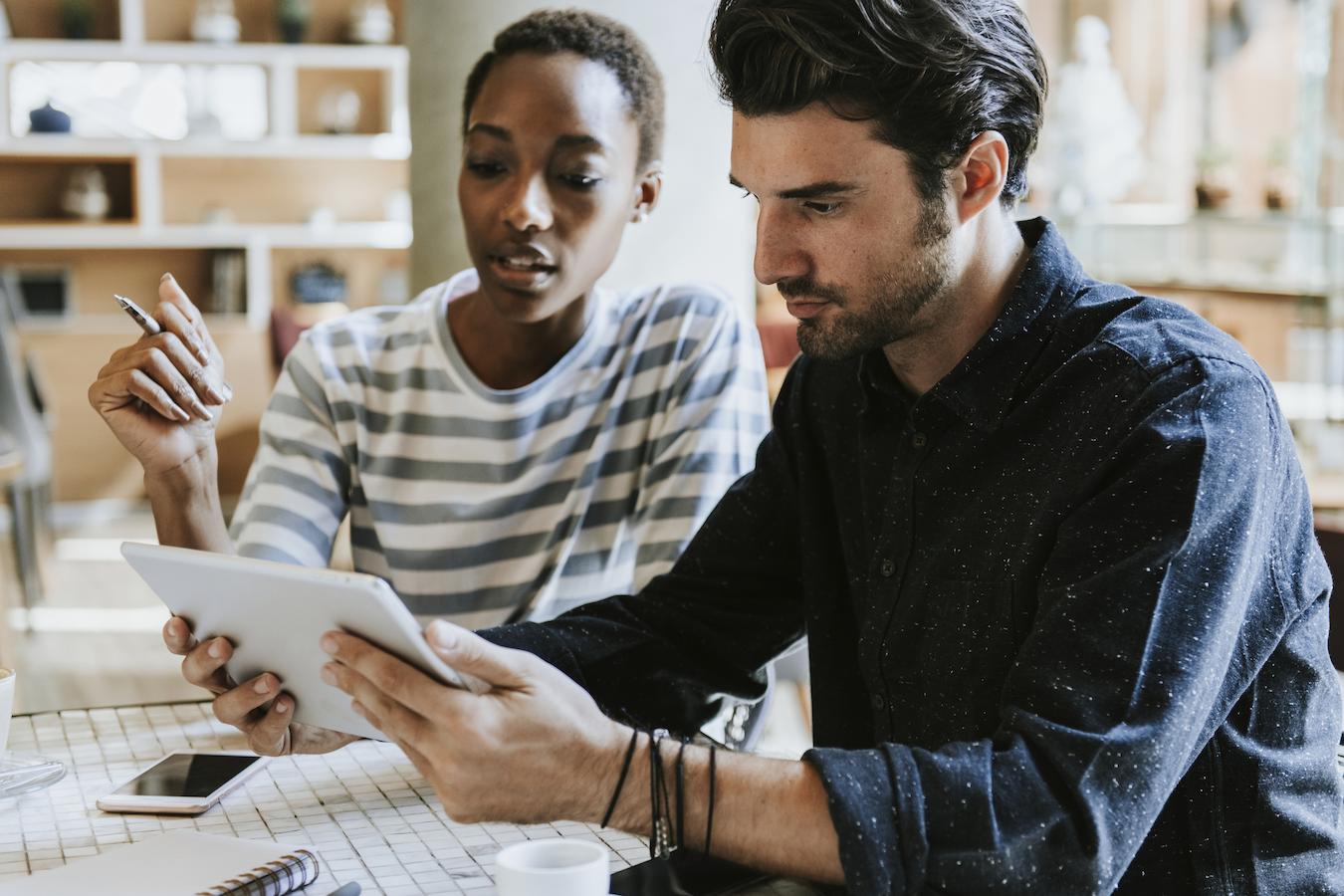 Two co workers sit at a desk and look at a tablet together